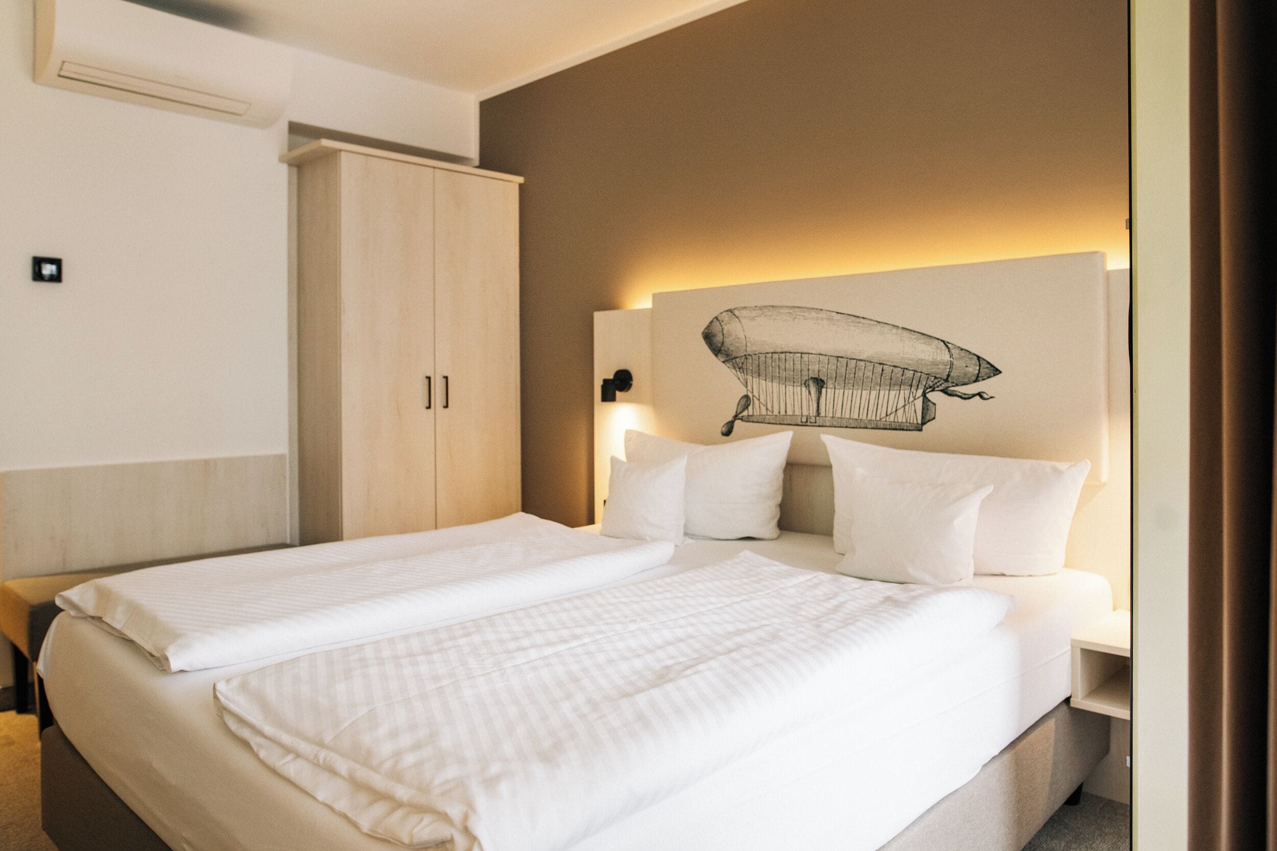 Una cama doble con sábanas blancas, un armario de madera y una lámpara fijada a la pared.