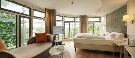 Een moderne hotelkamer met een sofa, een bed, een klein tafeltje met waterflesjes, een televisie en grote ramen met uitzicht op groen.