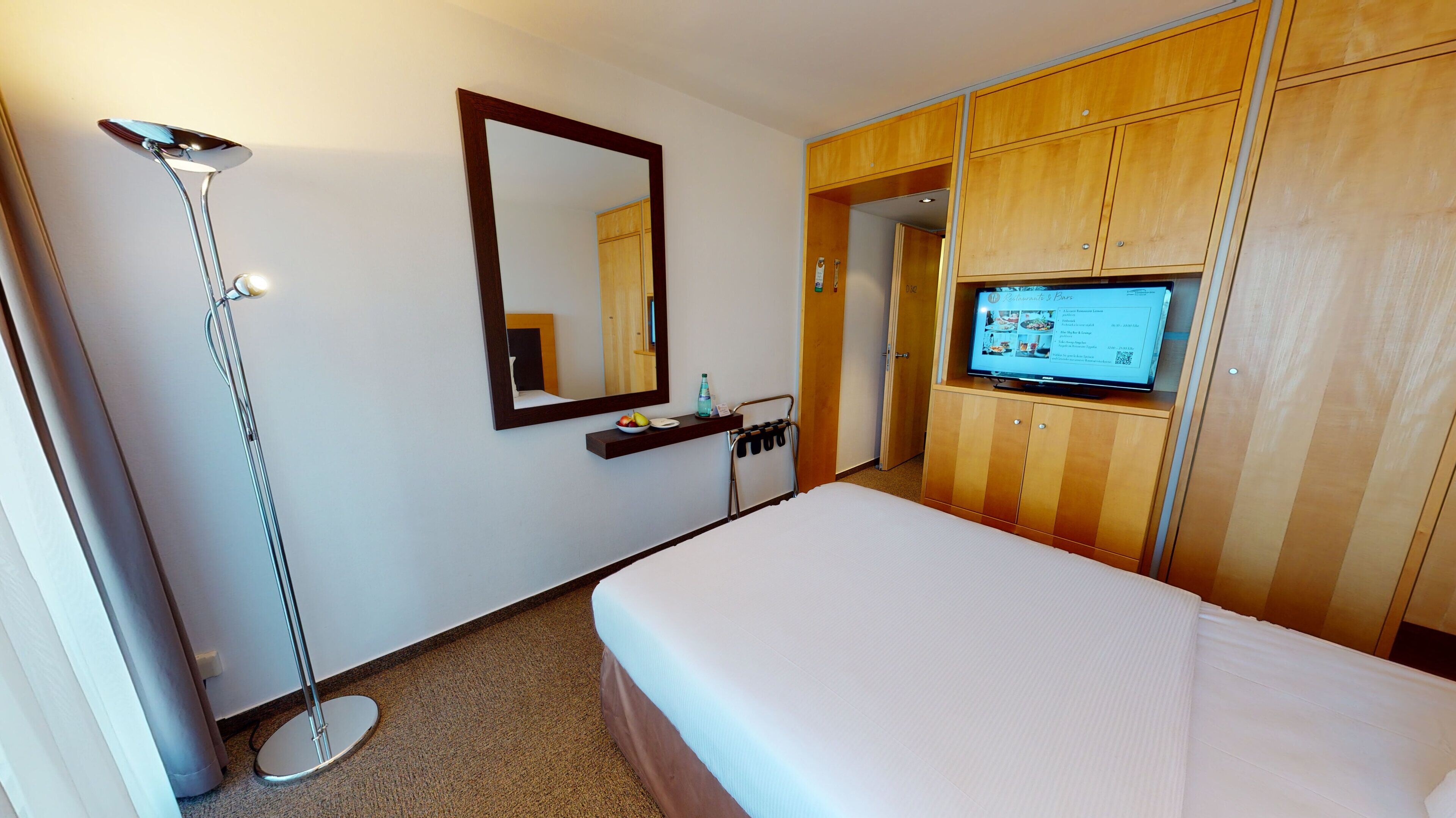 Ein Hotelzimmer mit einem Bett, einem an einem Holzkasten montierten Fernseher, einem Spiegel an der Wand und einer Stehlampe.
