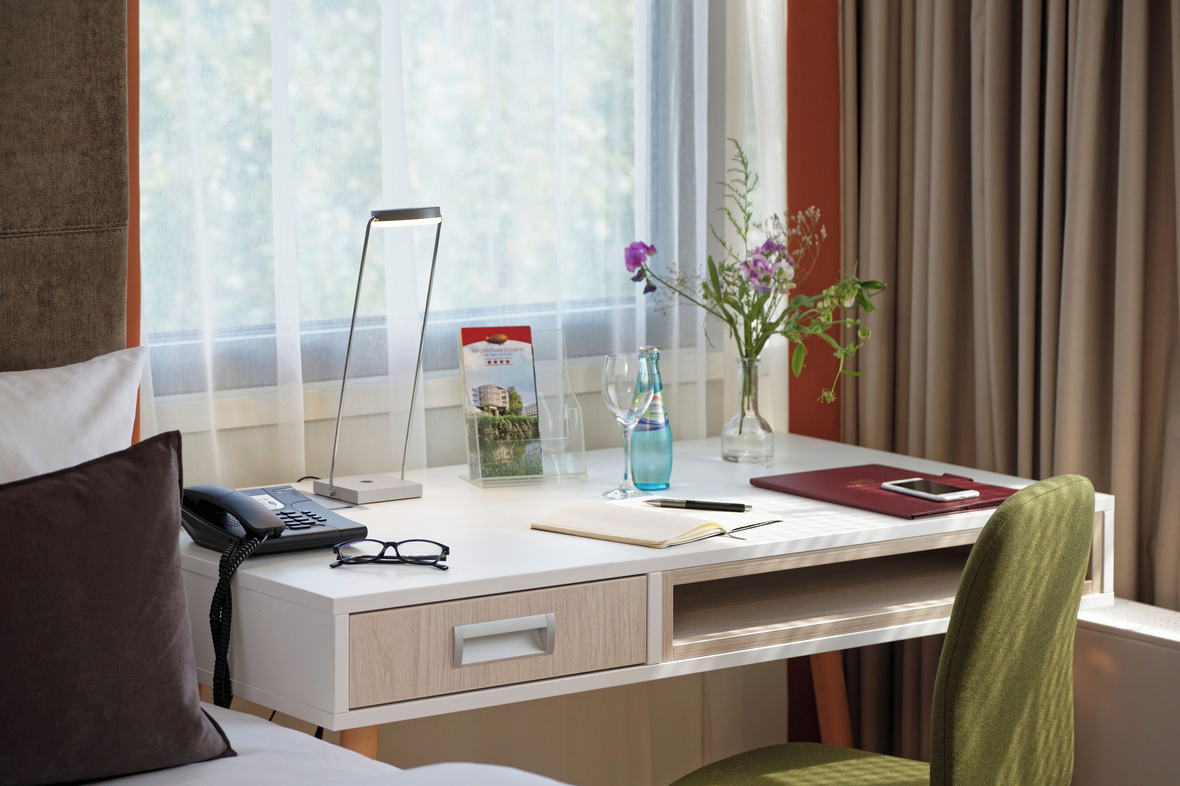 A hotel room with a desk, a phone, a notepad, a pen, a smartphone, a vase with flowers, and a window with curtains.
