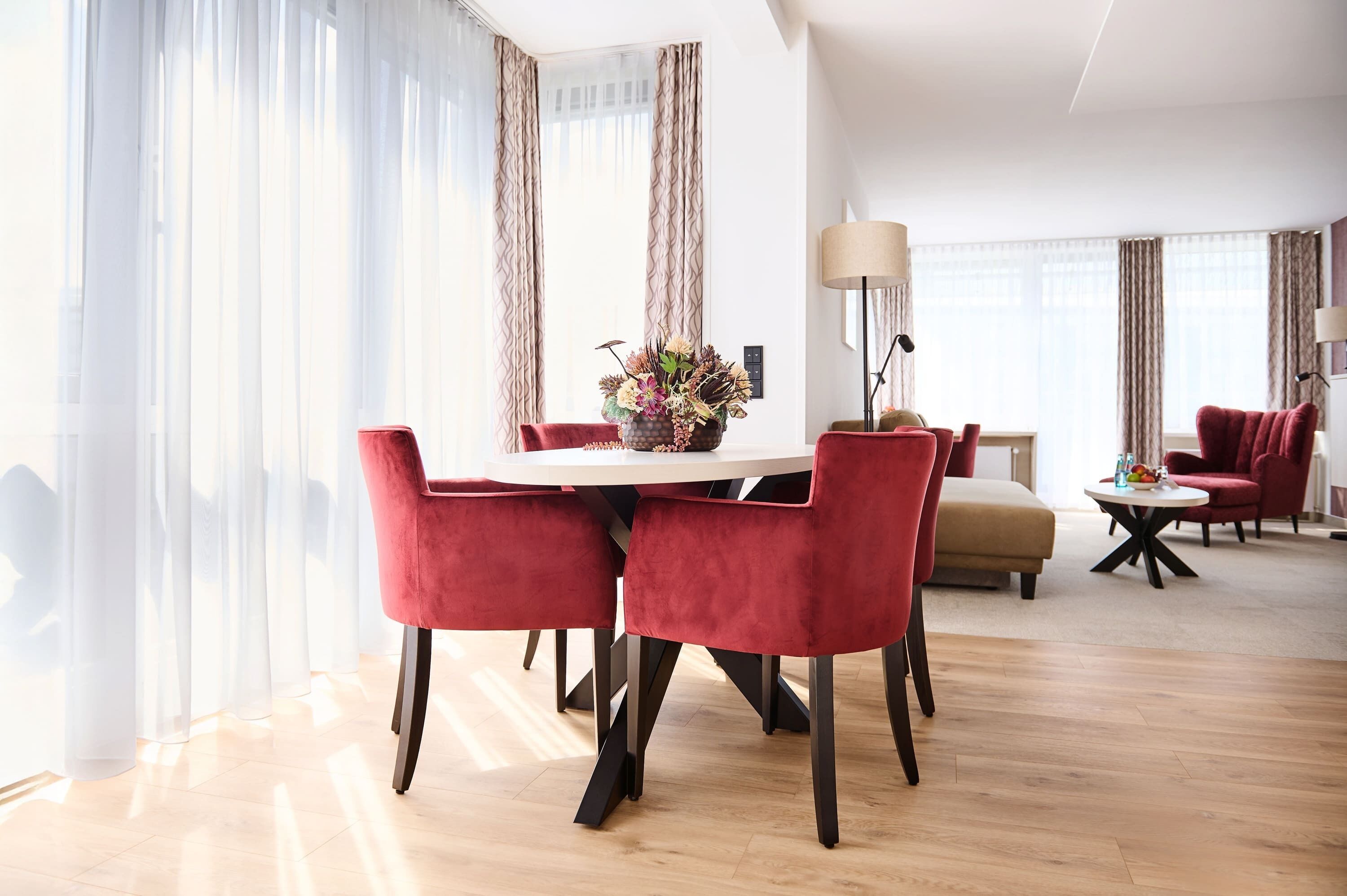 A modern dining area with a round table and red upholstered chairs, a vase of flowers, and a lamp on a side table.