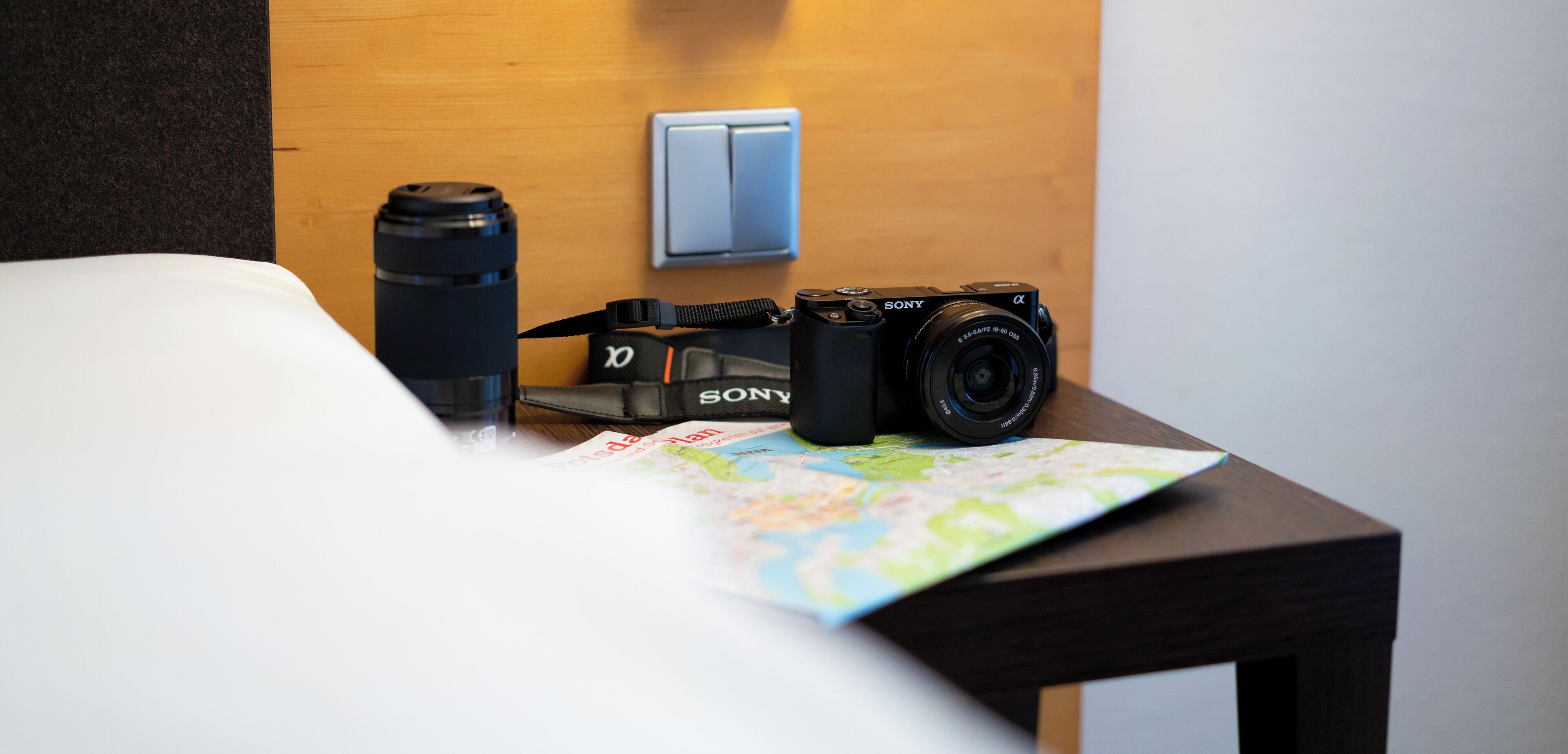 A neatly made bed with a map and a Sony camera on a wooden nightstand.