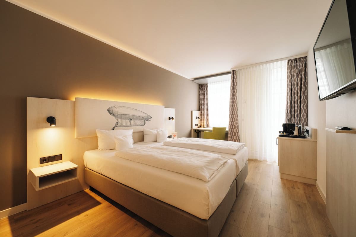 Una habitación de hotel moderna con una cama grande, mesitas de noche, un televisor de pantalla plana y vistas a la ciudad a través de amplias ventanas.