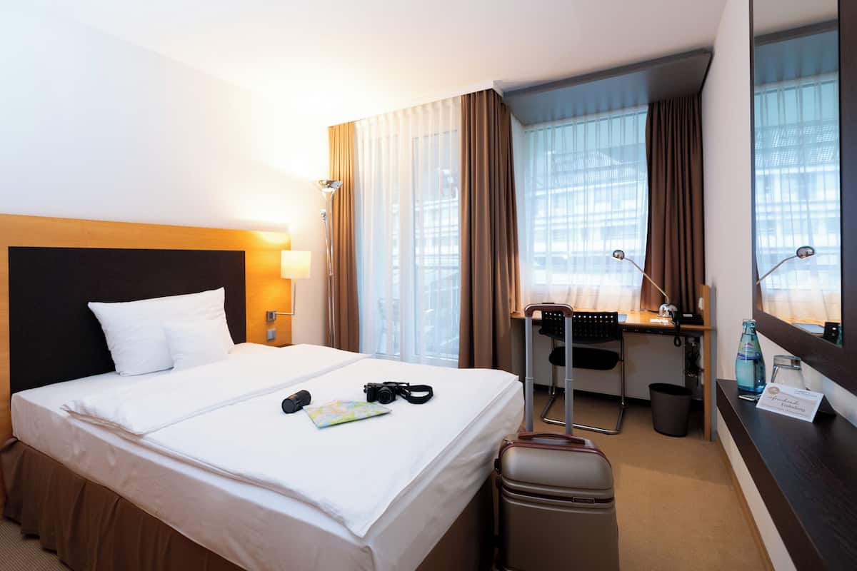 Una habitación de hotel con cama, escritorio, silla, maleta y lámpara de escritorio.