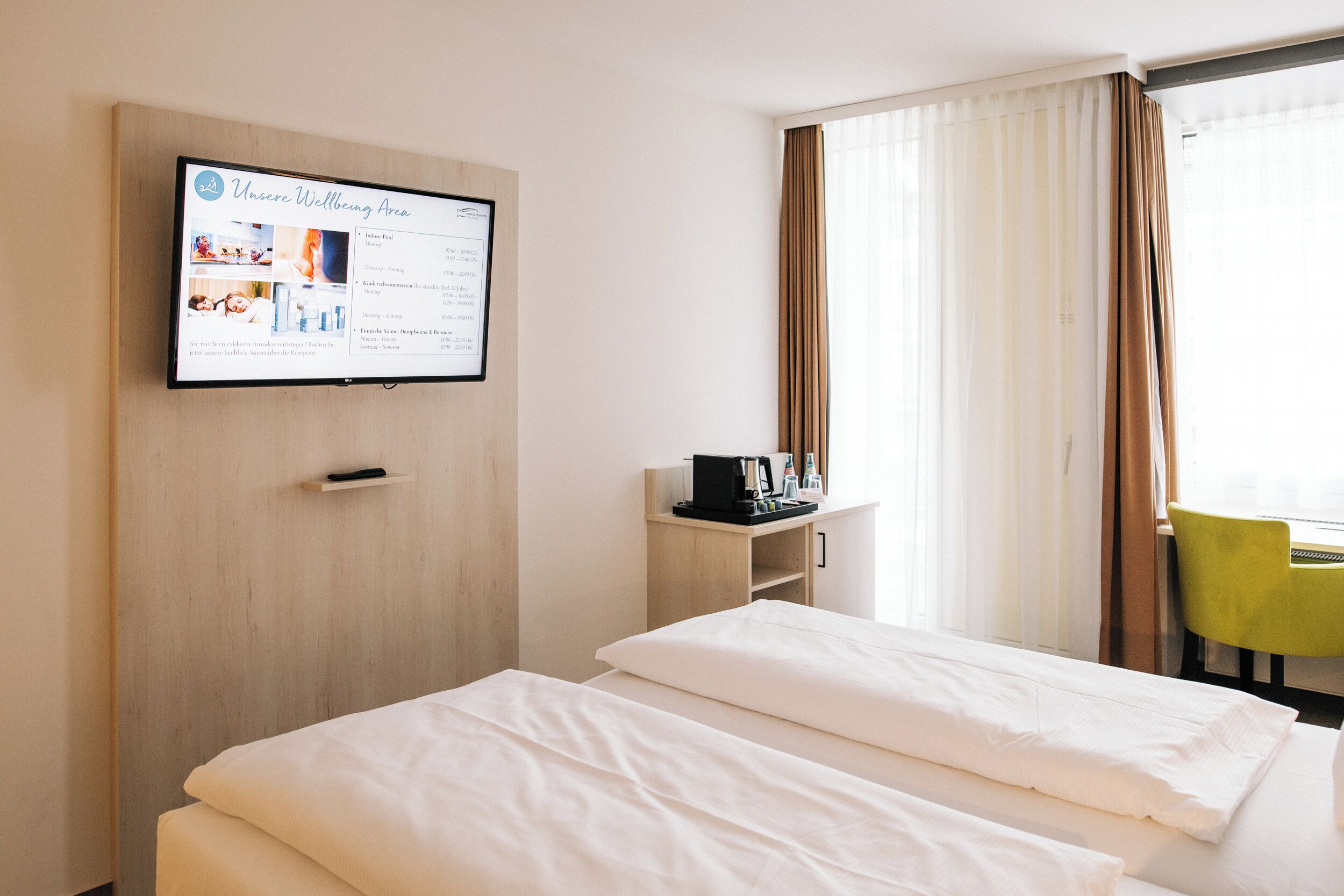 A hotel room with two beds, a mounted TV displaying a wellness area, a small desk with a coffee machine, and a view of a green chair outside.