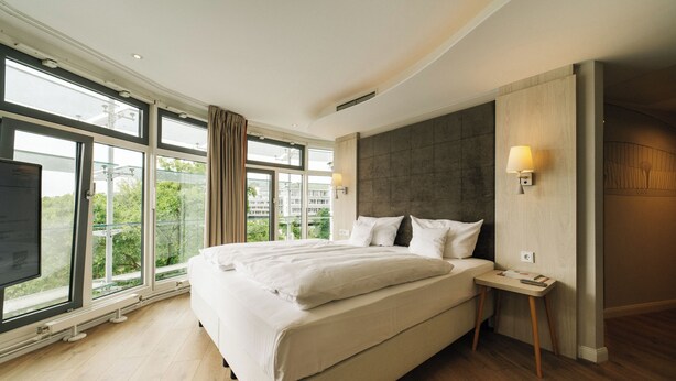 Een moderne hotelkamer met een groot bed, een nachtkastje en uitzicht door grote ramen.