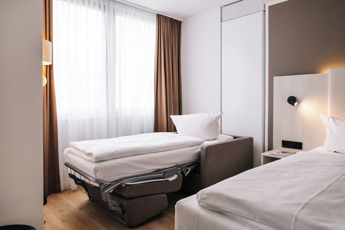 Habitación de hotel con dos camas, un escritorio, una silla, una lámpara y una ventana con cortinas.
