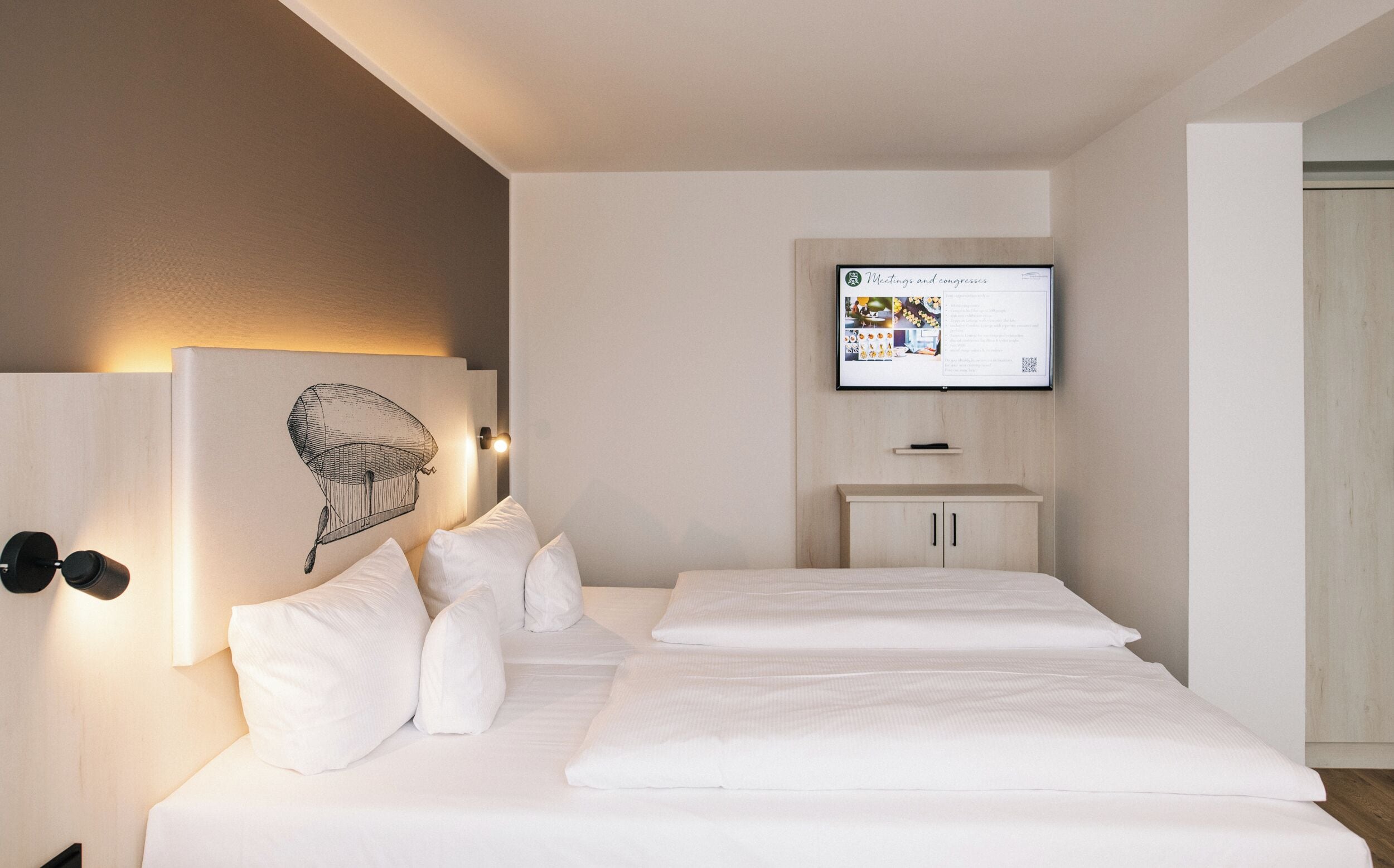 A hotel room with a bed, white bedding, a wall-mounted TV displaying a menu, and a light-colored wardrobe.