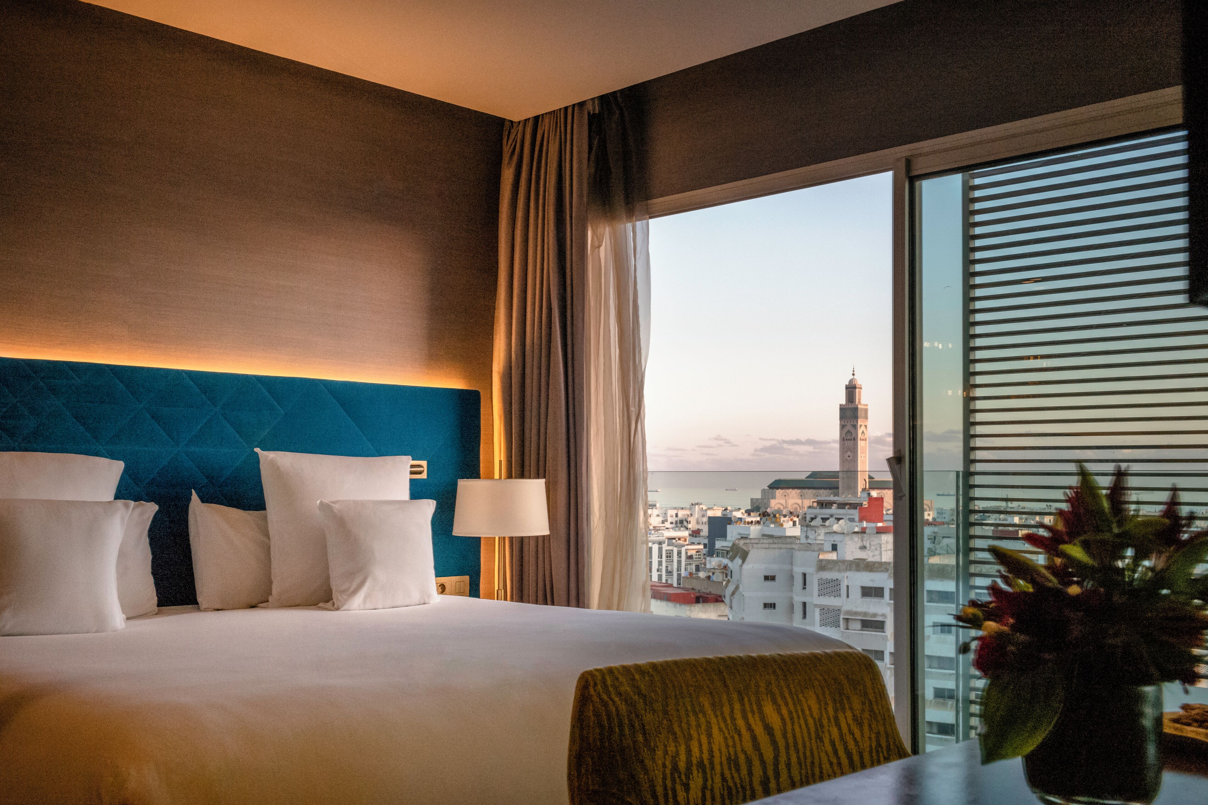 A hotel room with a large bed, a view of a cityscape, and a window with blinds.