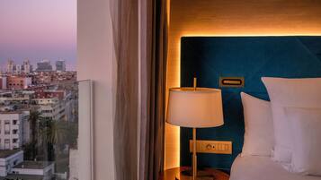 Une chambre d’hôtel avec vue sur la ville, un lit avec du linge de maison blanc, une table de chevet avec une lampe et une tête de lit bleue.