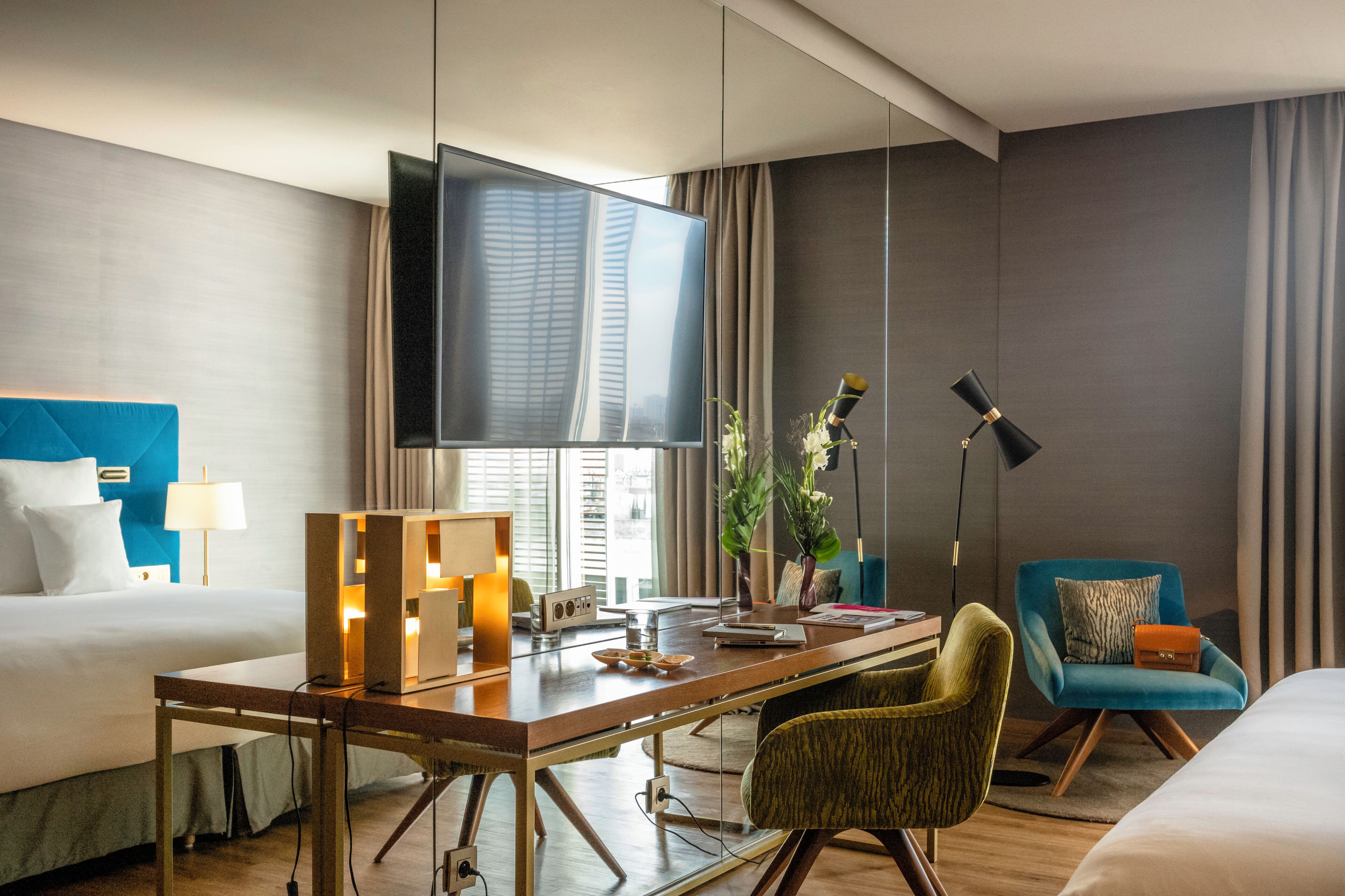 A hotel room with a bed, desk, TV, lamp, chair, and potted plants.