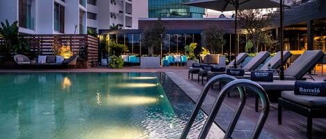 Zona de piscina con sillones de descanso, una piscina y un edificio al fondo.