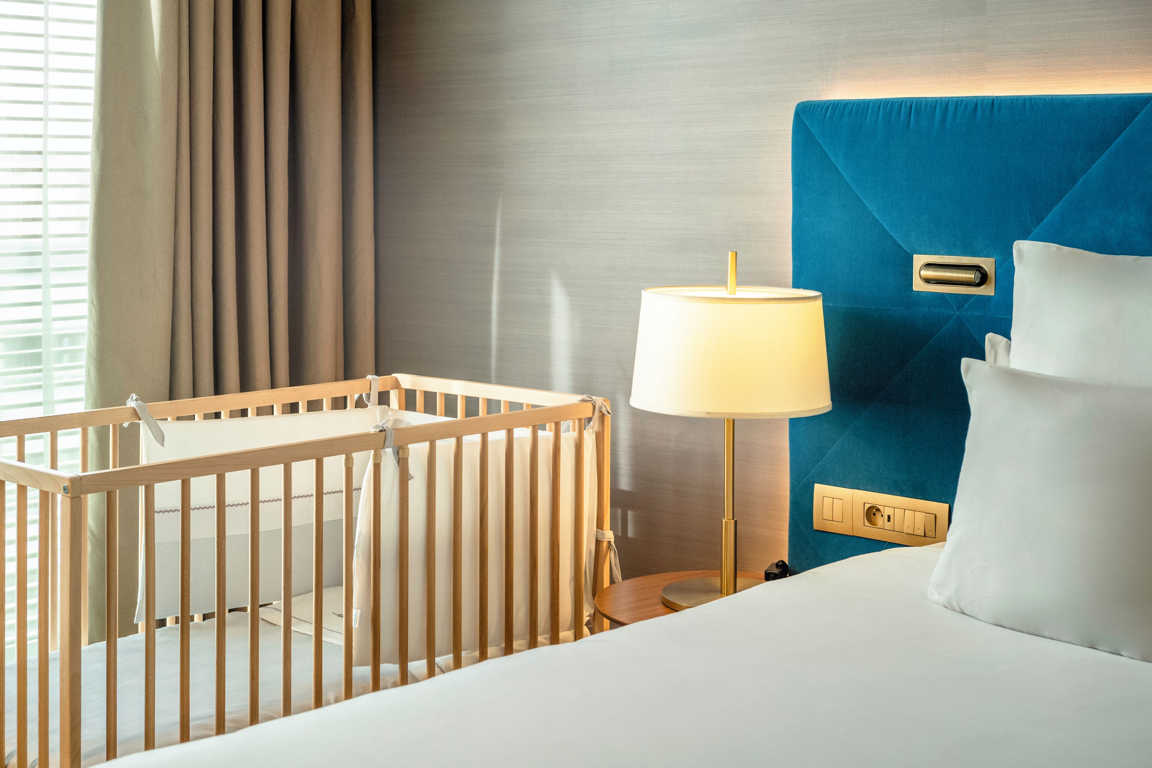 Une chambre d’hôtel avec un berceau, une lampe de chevet, un lit avec une literie blanche et une tête de lit bleue.