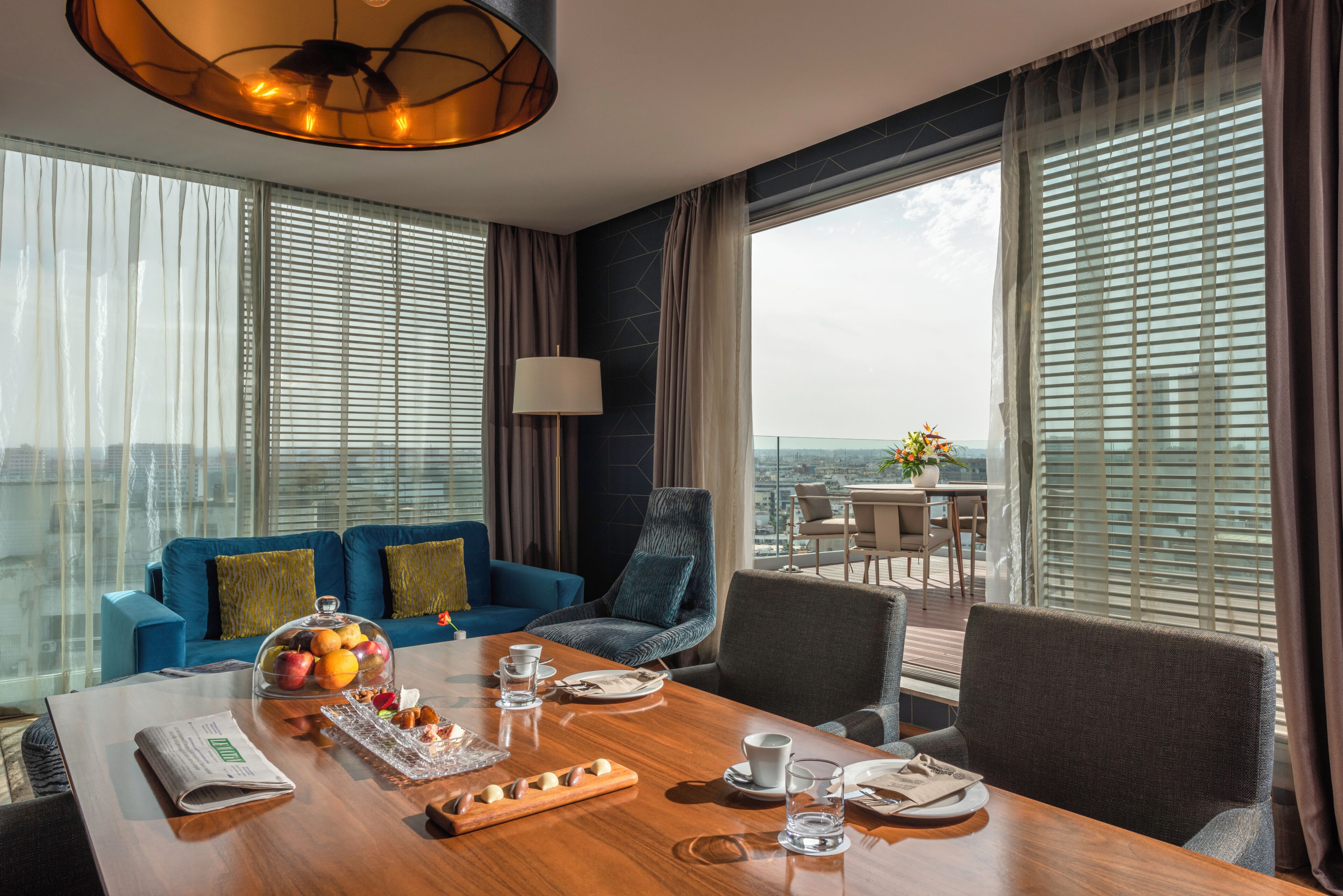 A dining area with a table set for two, a blue sofa, a chair, and a view of the city through large windows.