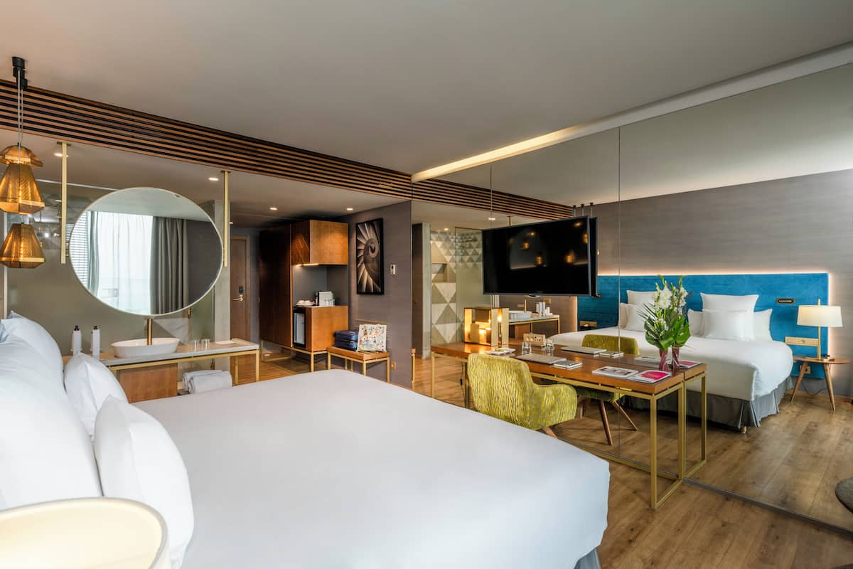 A modern hotel room with a large bed, a desk, a chair, a TV, and a bathroom with a mirror and sink.