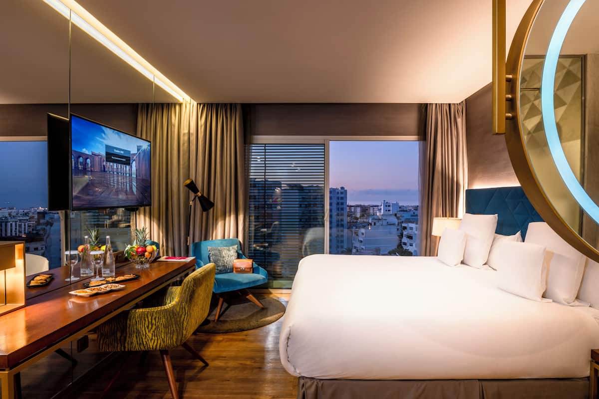 A hotel room with a large bed, a desk with a flat-screen TV, a dining area, and a view of the cityscape.