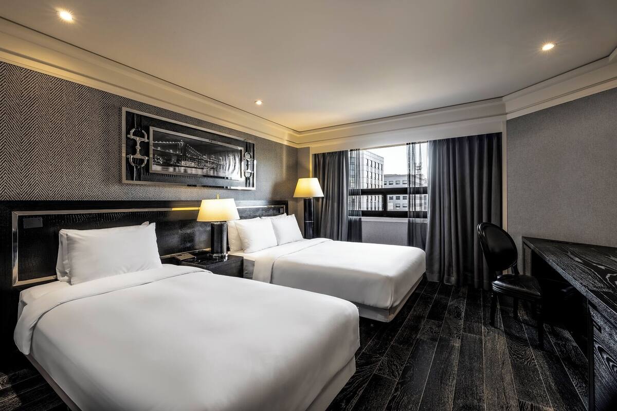 Une chambre d’hôtel avec deux lits, un bureau, une chaise, une fenêtre avec des rideaux et une œuvre d’art accrochée au mur.