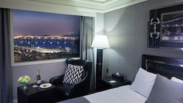 A hotel room with a large window offering a nighttime city view, a bed with white linens, a black chair with a patterned pillow, a bedside table with a bottle and glass, and a lamp.