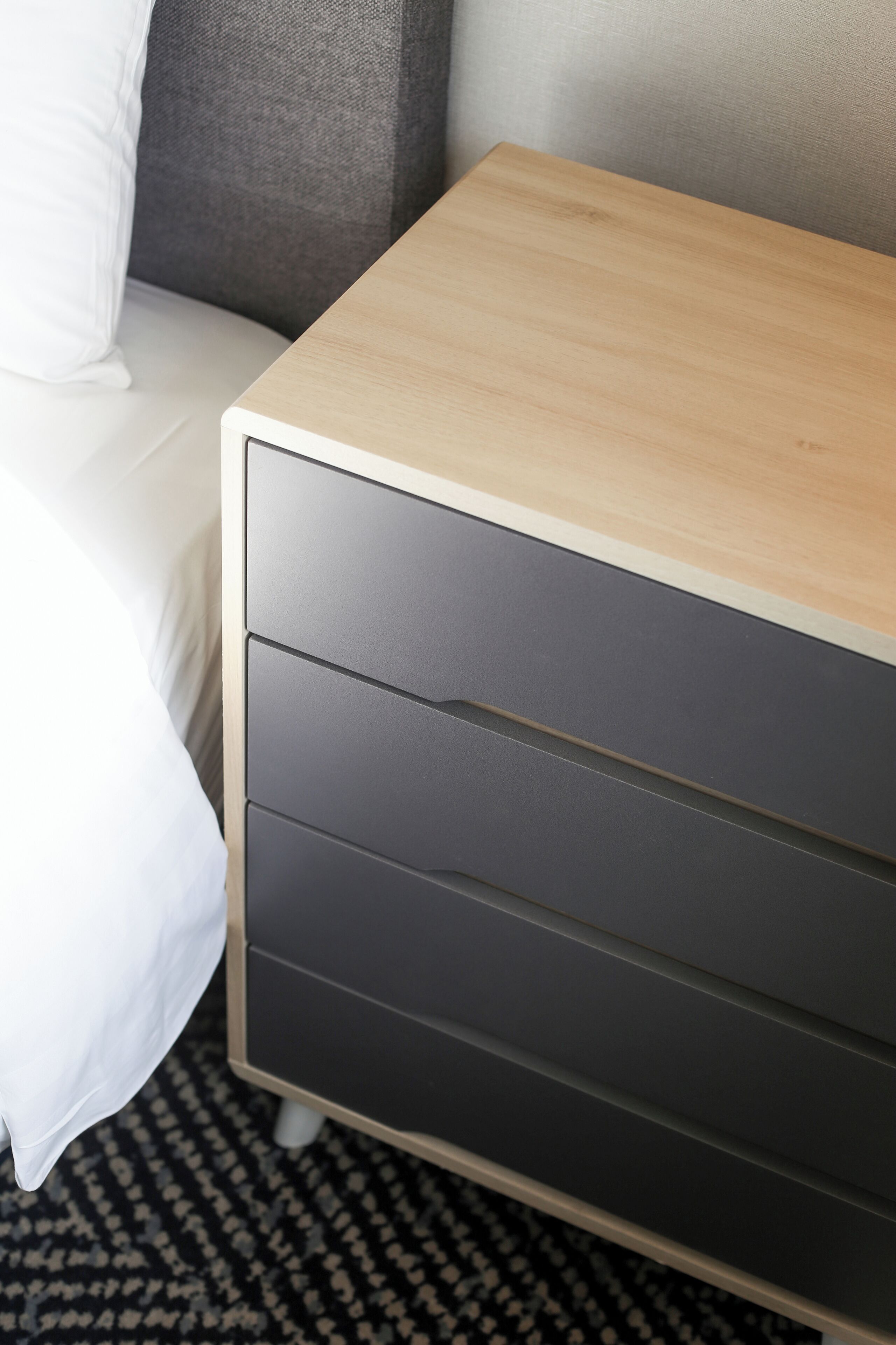Una cama con sábanas blancas, un almohadón gris y una mesita de noche de madera con cajón.