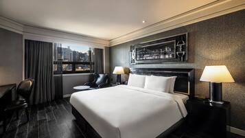 A modern hotel room with a large bed, two black leather chairs, a small round table, and a city view through the window.