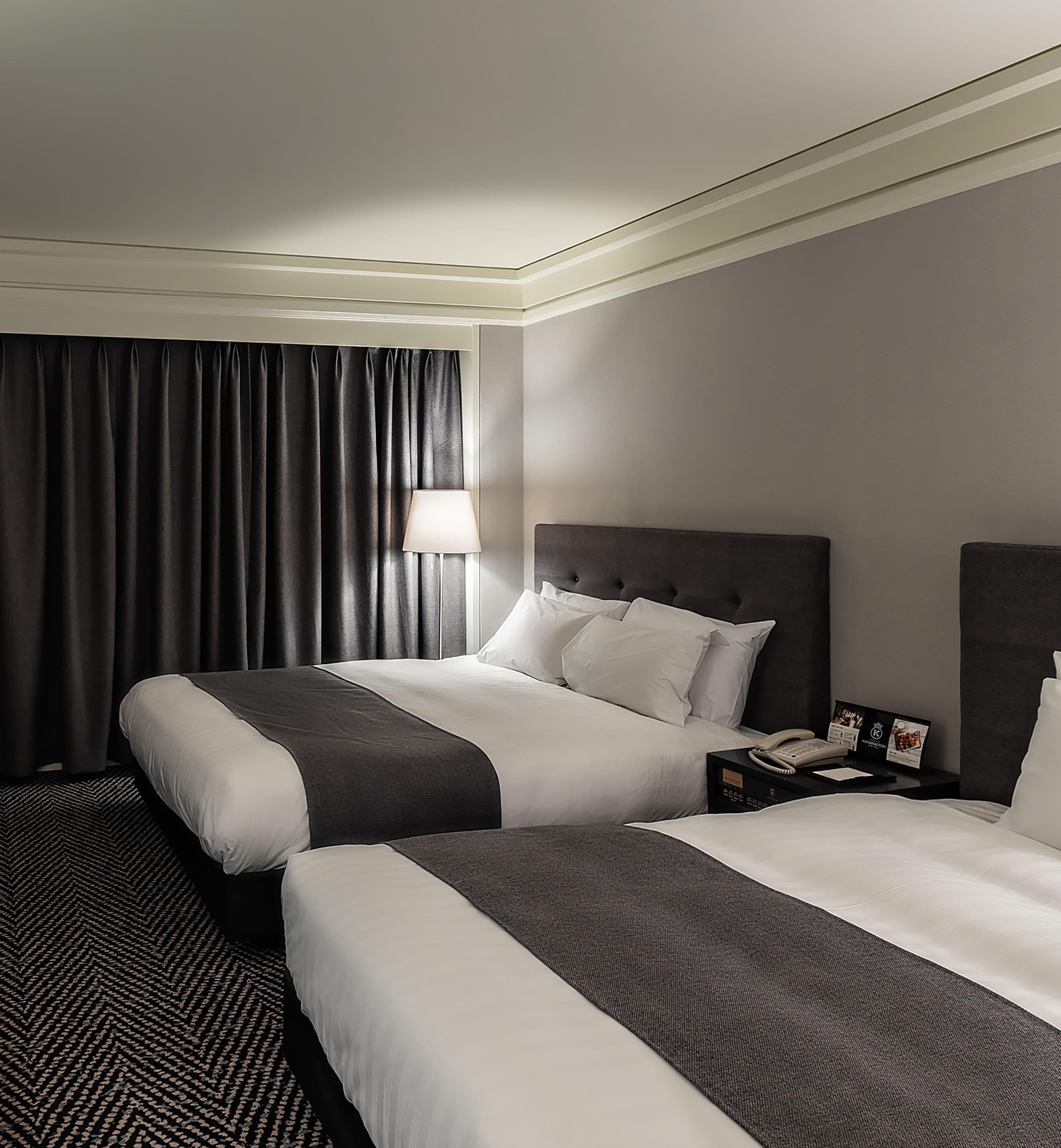 A hotel room with two beds, a nightstand with a telephone, and a lamp.