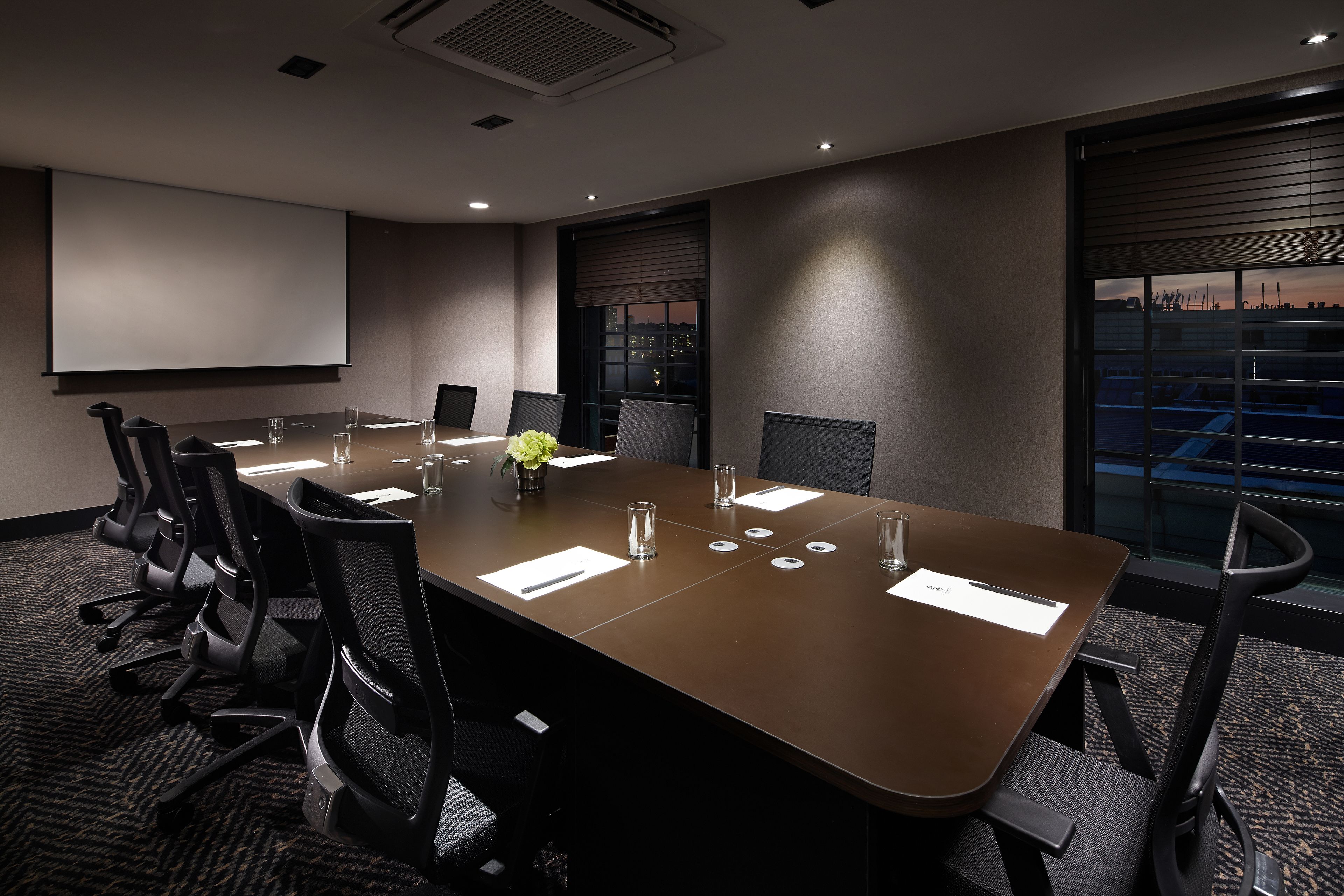 A conference room with a long table, chairs, a projector screen, and a window with a city view.
