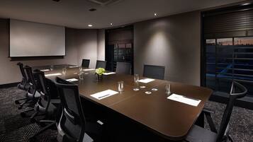 A conference room with a long table, chairs, a projector screen, and a window with a city view.