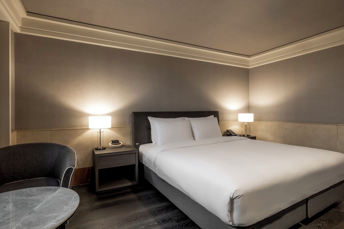 A hotel room with a large bed, two bedside tables with lamps, a chair, and a small table.