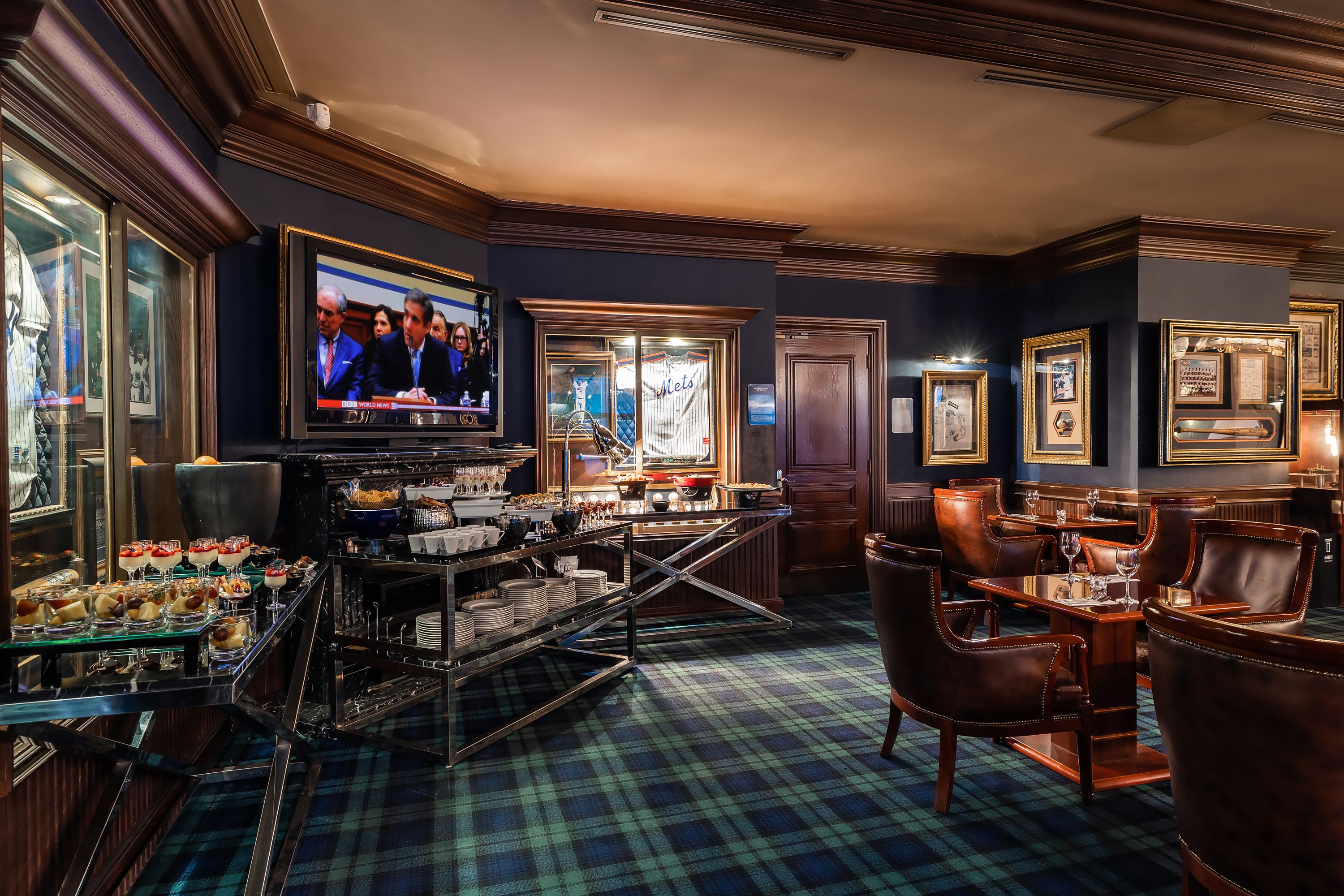 A bar area with a television displaying a news channel, a buffet with various dishes, and a seating area with leather chairs and a plaid carpet.