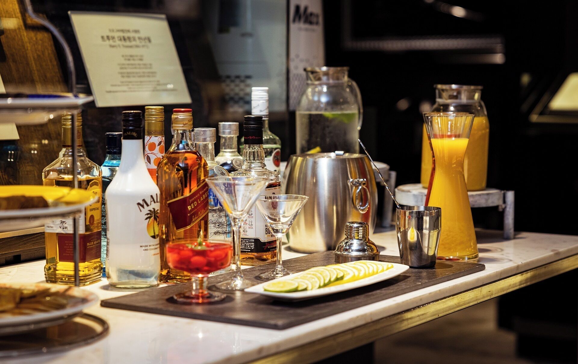 Un coin bar avec différentes bouteilles d’alcool, un verre à martini, un shaker à cocktails et un plateau contenant des tranches de citron.