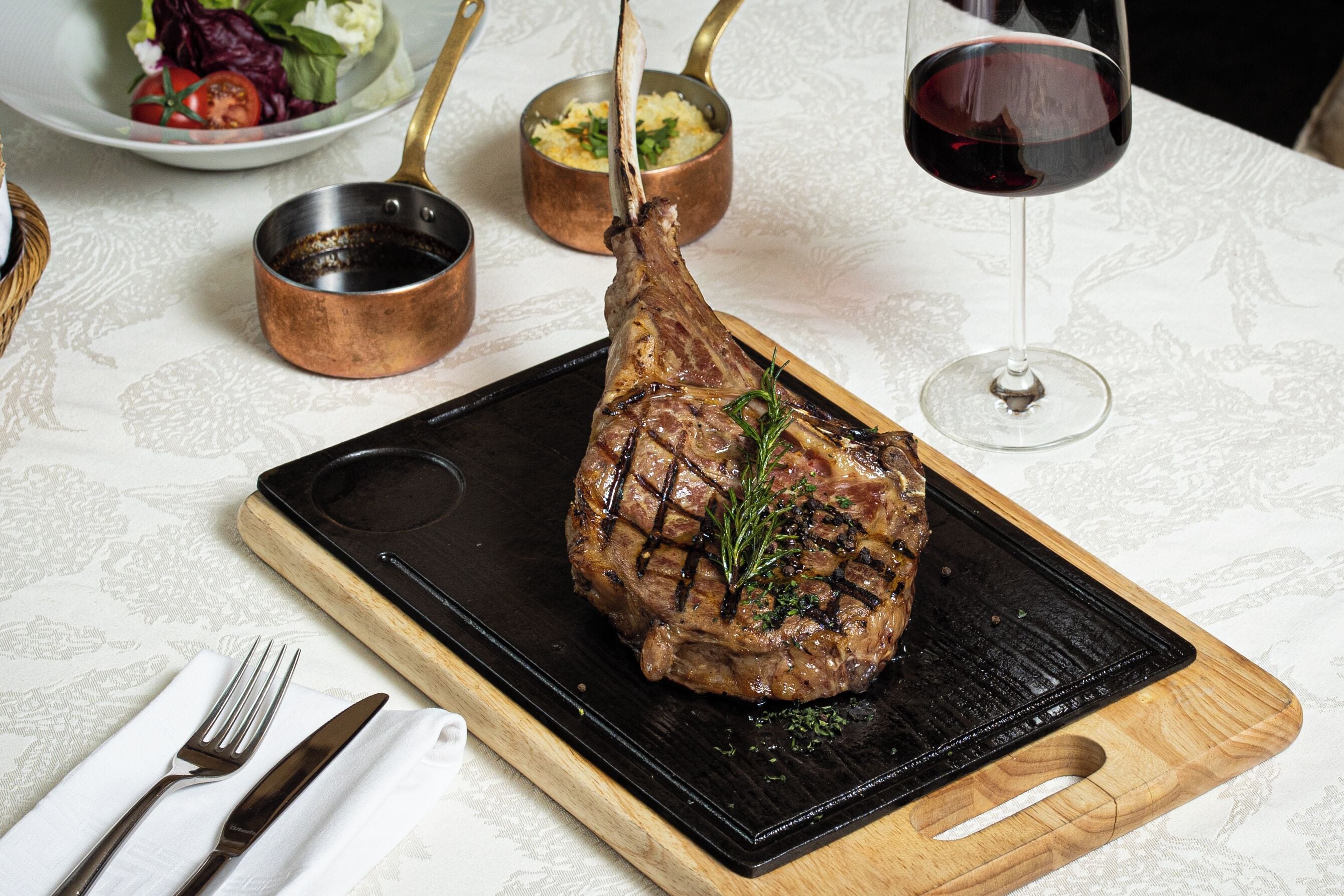 A roasted lamb leg on a wooden board with a black grill, accompanied by a glass of red wine and a bowl of salad.