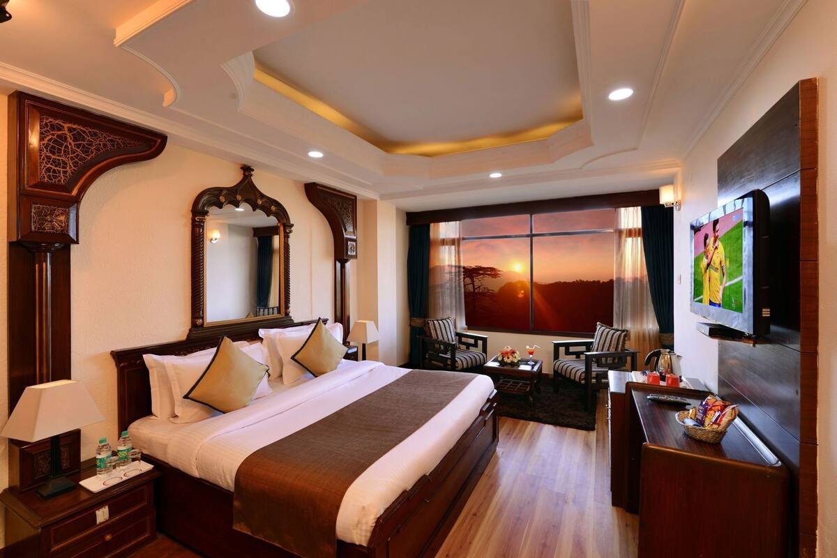 A hotel room with a large bed, a TV, a desk with a chair, a window with a sunset view, and a decorative mirror.