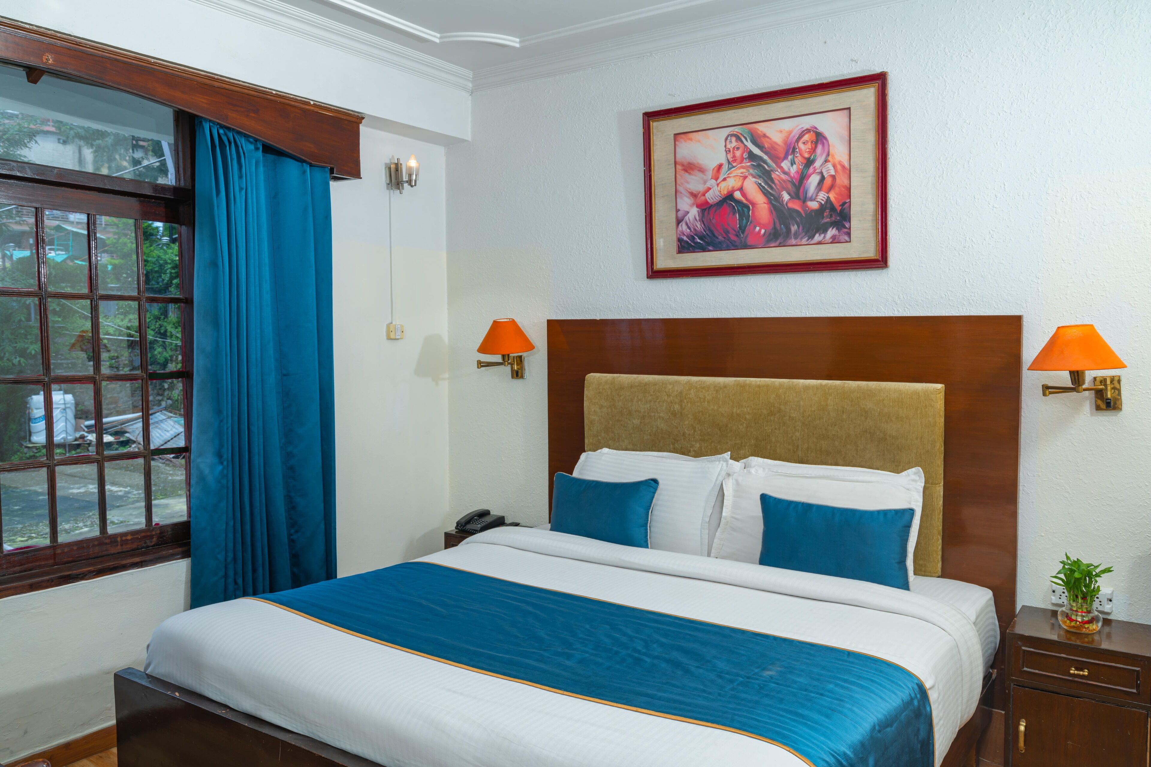 A hotel room with a large bed, blue curtains, a framed painting, and two wall-mounted lamps.