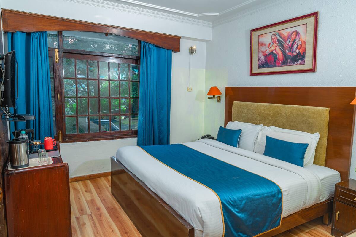 A hotel room with a large bed, a wooden headboard, a window with curtains, a painting on the wall, and a nightstand with a lamp.