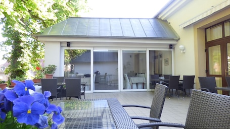 An outdoor patio with a glass door leading to an interior dining area, surrounded by wicker furniture and potted plants.