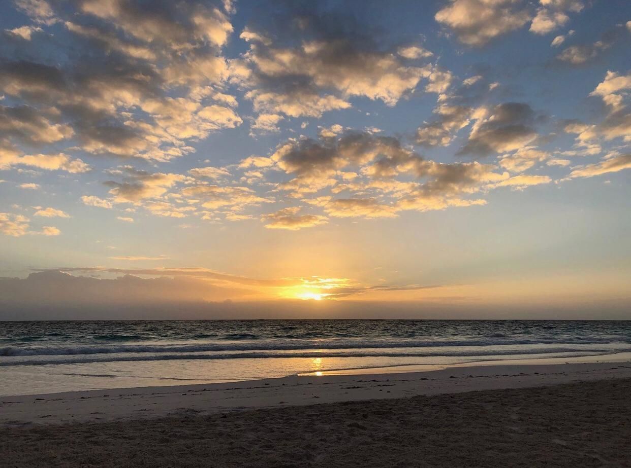Ein Strand mit untergehender Sonne am Horizont, die eine Silhouette aus Wolken am Himmel wirft.