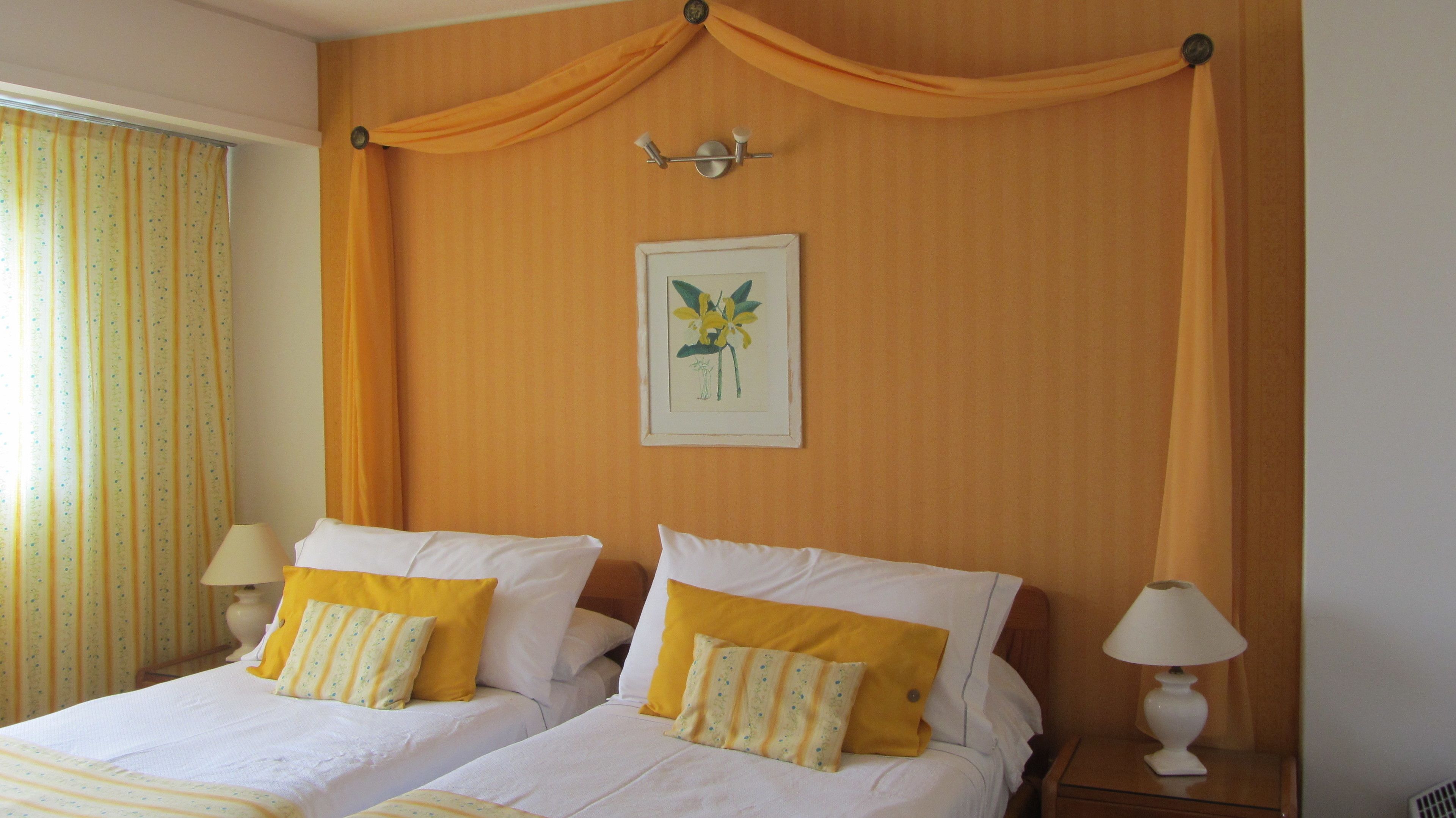 A double bed with yellow and white pillows, a framed floral artwork on the wall, and a window with curtains.