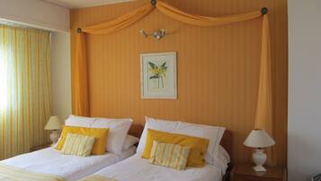 A double bed with yellow and white pillows, a framed floral artwork on the wall, and a window with curtains.