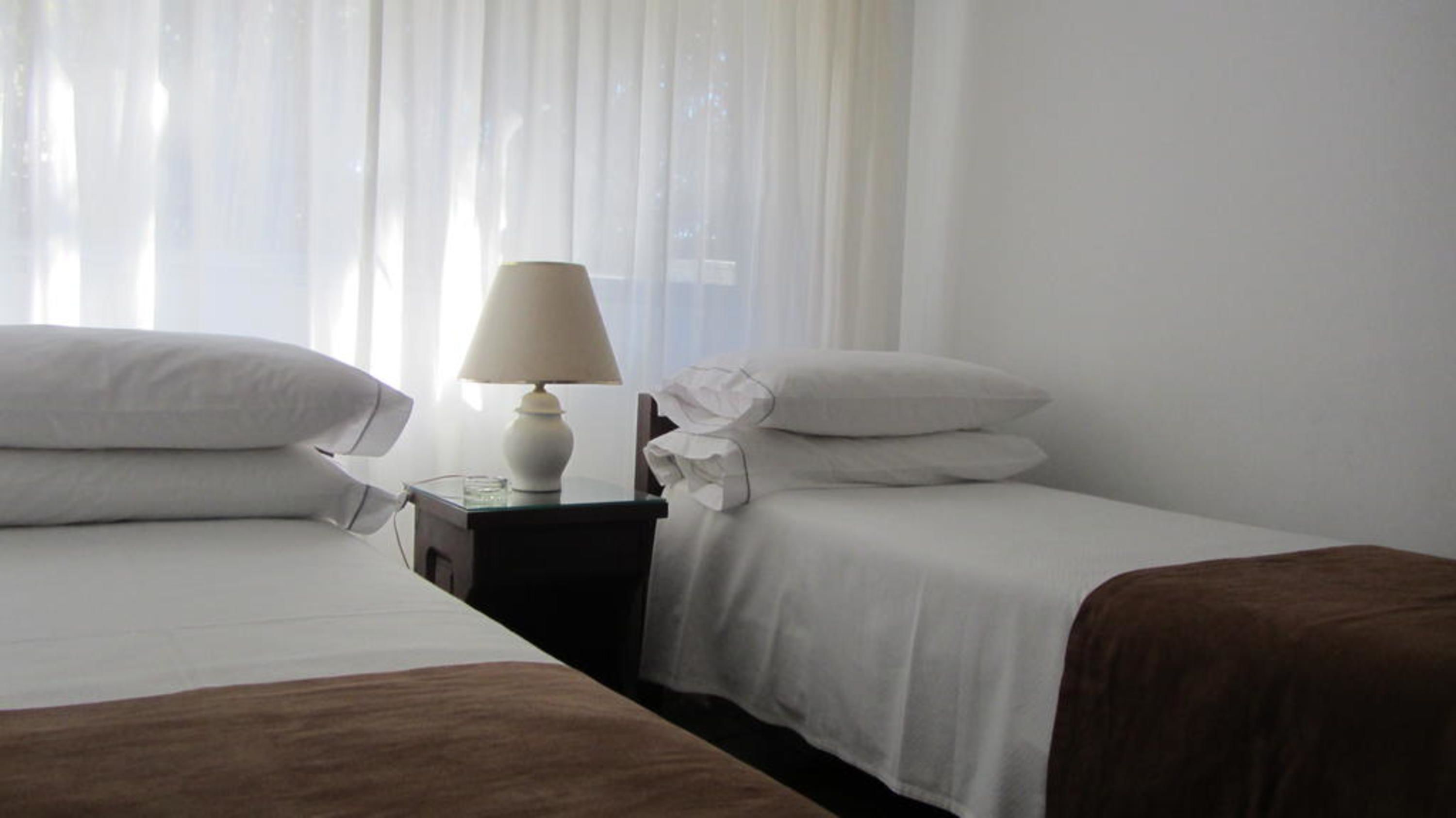 Habitació d'hotel amb dos llits, una nitòria amb una làmpada i cortines transparents.