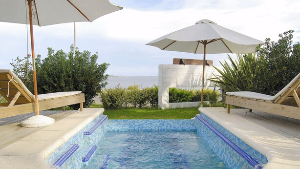 Un jacuzzi con borde de azulejos, rodeado de sillones de descanso y sombrillas, con vista al mar de fondo.