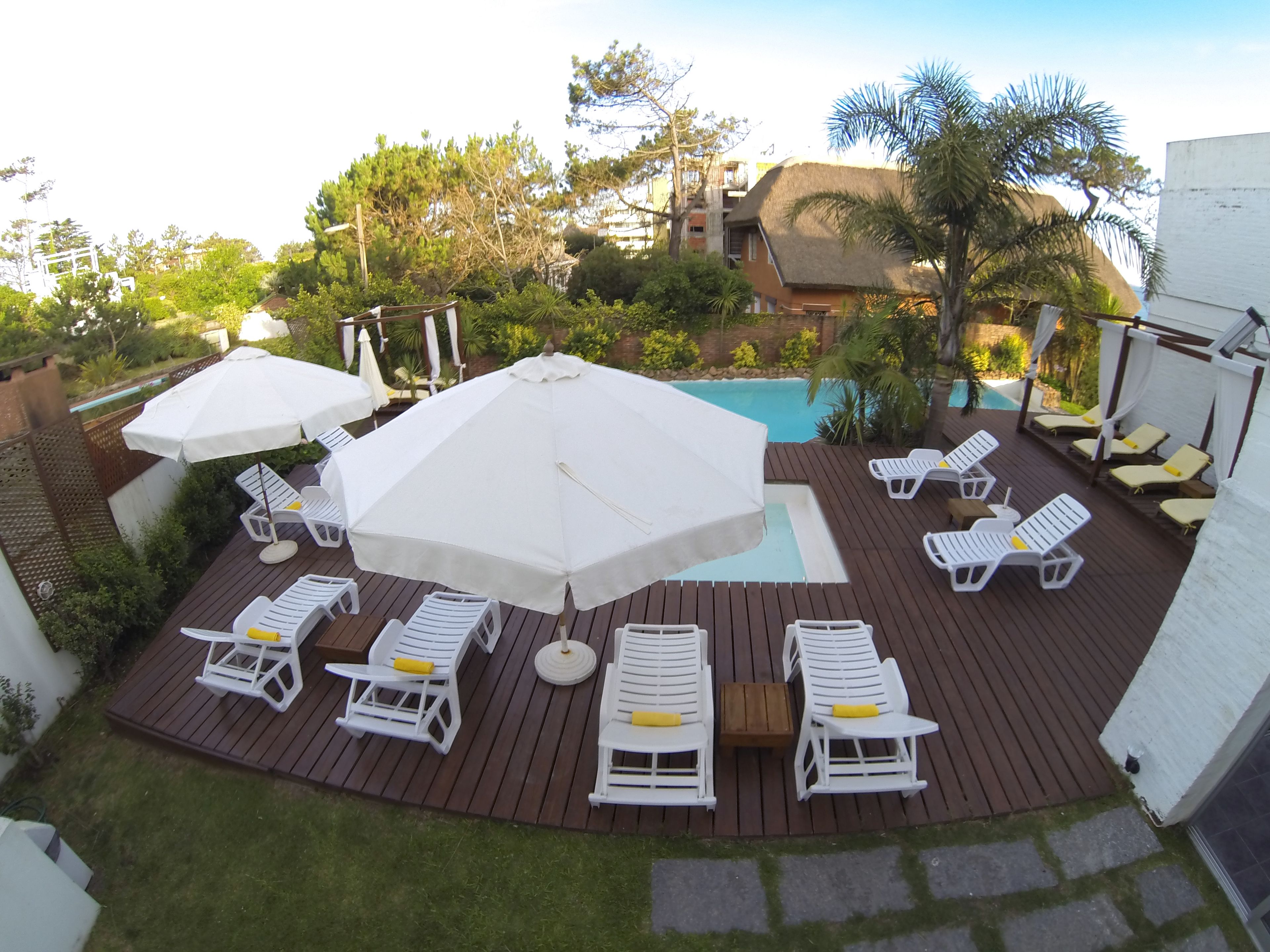 Una terraza de madera con sillones de descanso blancos y sombrillas, una piscina y vegetación circundante.