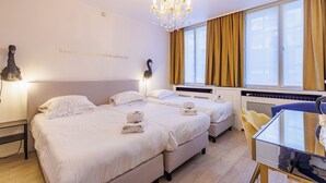 Habitación de hotel con dos camas, un candelabro, un ventanal grande con cortinas y una pequeña mesa con una silla.