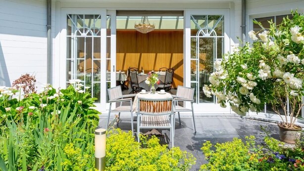 Un patio con una mesa y sillas, rodeado de un jardín con diversas plantas y flores.