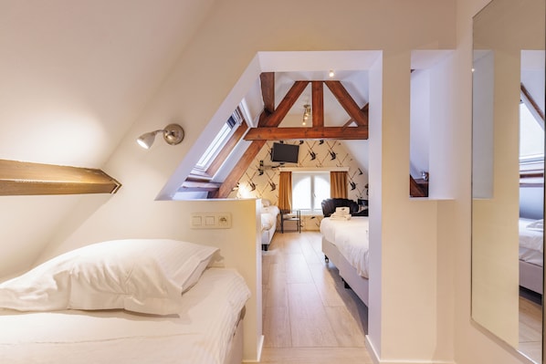 A bedroom with a sloped ceiling, wooden beams, a TV mounted on the wall, and a window providing natural light. - The Black Swan Hotel (Bruges)