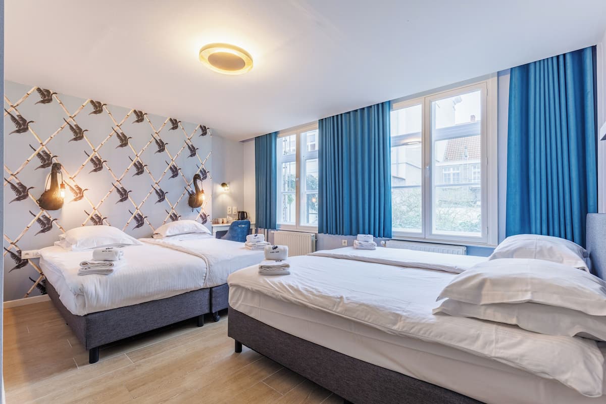 A hotel room with two beds, a patterned wall, a large window with blue curtains, and a ceiling light.