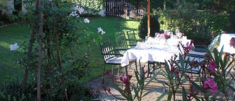 An outdoor dining area with a table set for two under a green umbrella, surrounded by lush greenery and flowering plants.