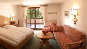 A hotel room with a bed, a wooden nightstand with a lamp, a small table with a vase, a sofa, and a window with curtains. - Hotel Gasthof Moosleitner (Freilassing)