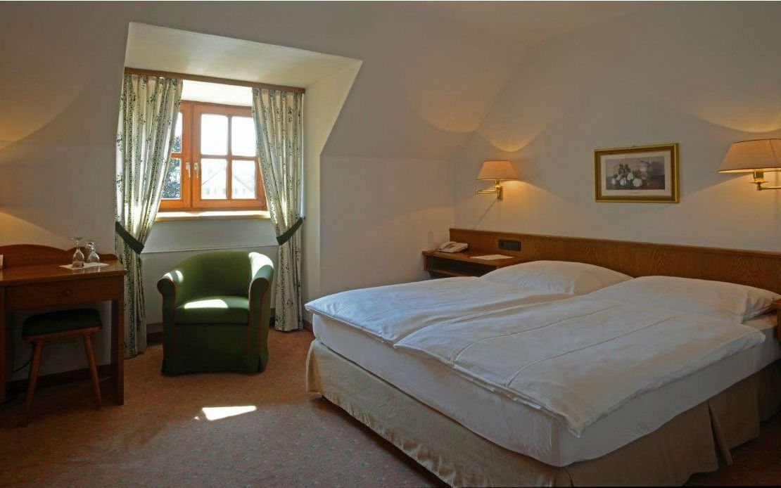 A hotel room with a bed, a desk, a chair, a window with curtains, and two wall-mounted lamps.