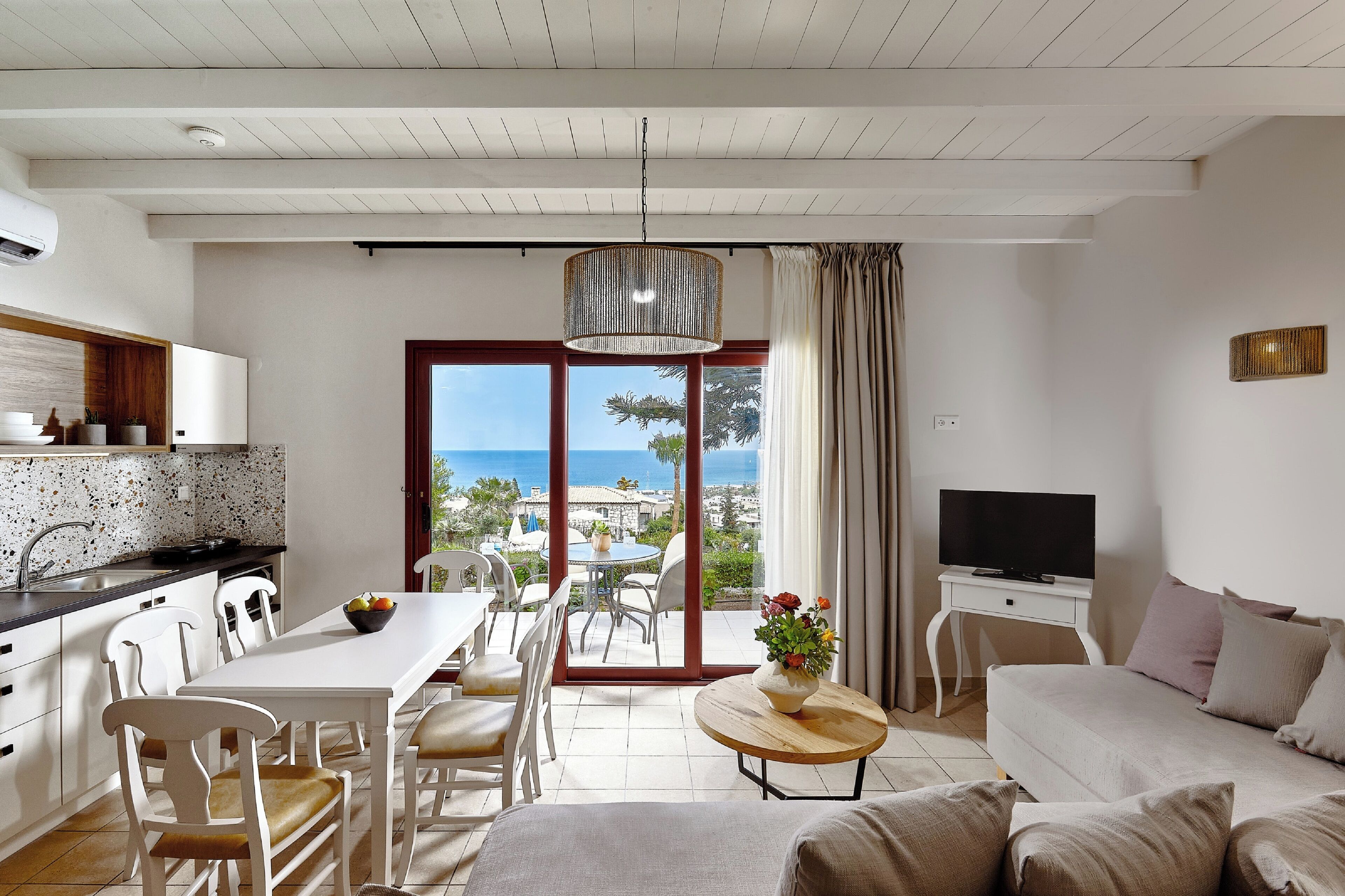Ein modernes Wohnzimmer mit Sofa, einem Esstisch mit Stühlen, einem Fernseher auf einem Schrank und Meerblick durch große Fenster.