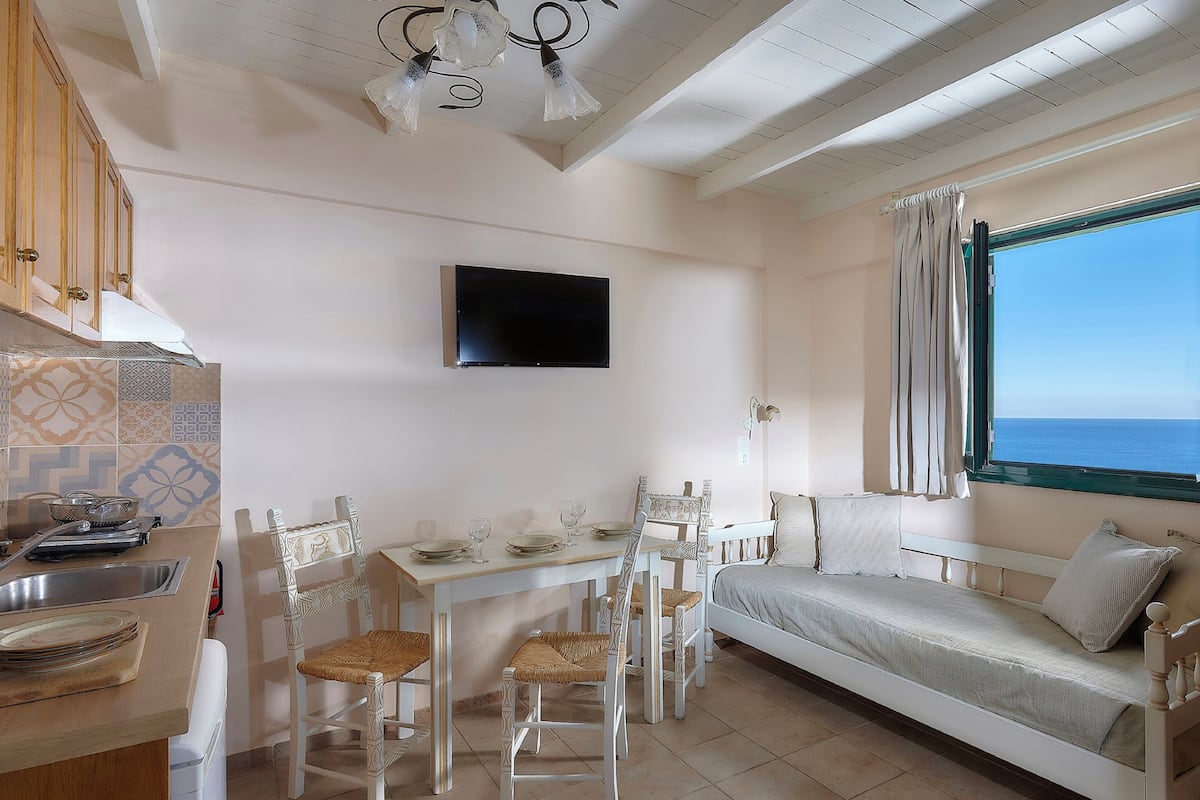 A compact room with a dining table, chairs, a sofa, a TV, and a view of the sea.